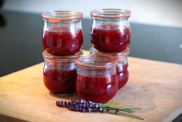 Five small glass jars filled with red jam are stacked on a wooden surface. In front of the jars, several sprigs of fresh lavender are placed as decoration.