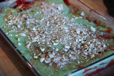 A glass baking dish filled with a dessert featuring a green creamy layer topped with crushed nuts, set on a wooden surface. Some red filling is visible around the edges of the dish.