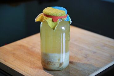 A glass jar filled with a cloudy, yellowish liquid sits on a wooden board. The jar is covered with a colorful cloth held by a rubber band, and sediment has settled at the bottom.