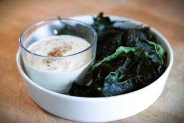 A white bowl with dark green kale chips and a glass cup of creamy dipping sauce topped with sesame seeds, placed on a light wooden surface.