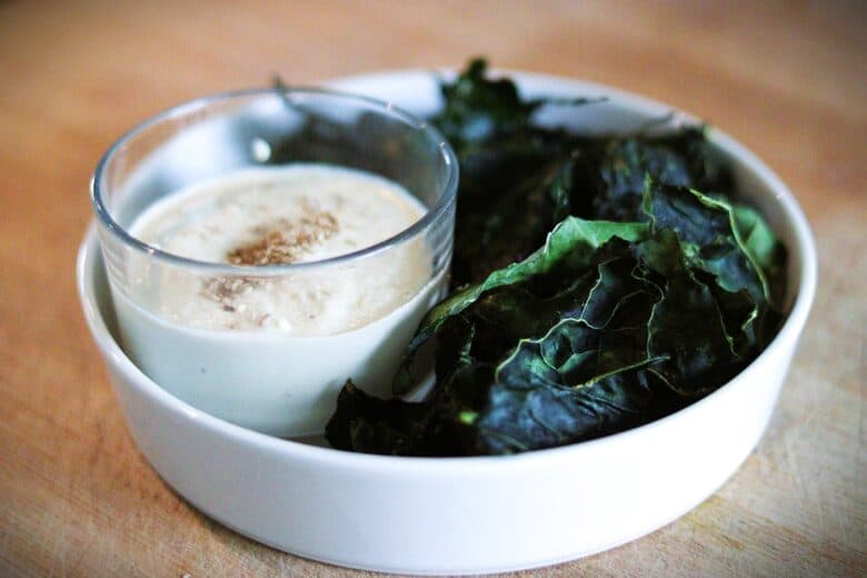 A white bowl with dark green kale chips and a glass cup of creamy dipping sauce topped with sesame seeds, placed on a light wooden surface.