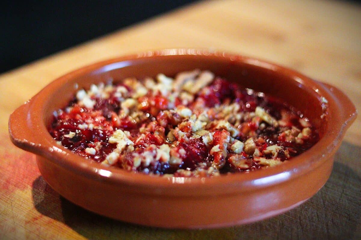 A close-up of a berry crumble dessert in a round brown ceramic dish, topped with chopped nuts, sitting on a wooden surface.