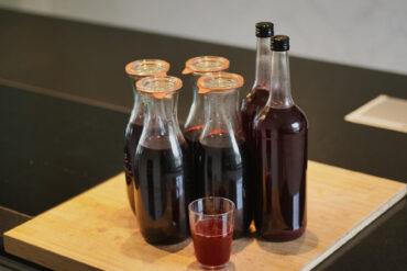 Five glass bottles filled with a dark red liquid are arranged on a wooden cutting board, with a small plastic cup containing the same liquid placed in front of them. The background is a dark countertop.
