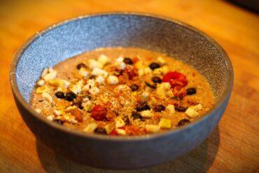 A gray bowl filled with oatmeal topped with diced apples, blueberries, nuts, seeds, and dried fruit, sitting on a wooden surface.