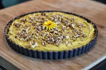 A round tart with a creamy yellow filling, topped with chopped nuts and a yellow flower in the center, sits on a wooden surface in a fluted metal pan.