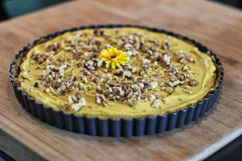 A round tart with a creamy yellow filling, topped with chopped nuts and a yellow flower in the center, sits on a wooden surface in a fluted metal pan.