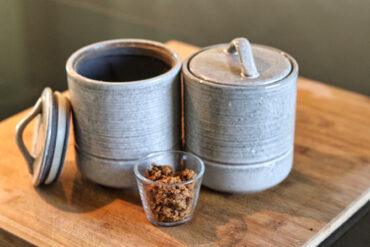 Two gray ceramic jars, one with a lid off and the other with a lid on, are on a wooden surface. In front of them is a small clear glass cup containing brown sugar or a similar granulated substance.
