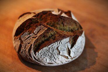 A round, rustic loaf of artisan bread with a dark, crusty top and a dusting of flour sits on a wooden surface.