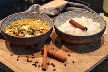 Two bowls on a wooden board: one with yellow curry, greens, and chicken; the other with white rice topped with a cinnamon stick. Extra cinnamon sticks and cloves are scattered nearby. A gray cloth is in the background.