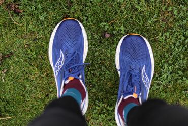 Person wearing blue athletic shoes and colorful socks with patterns, standing on green grass. The photo is taken from above, showing only the lower legs and feet.