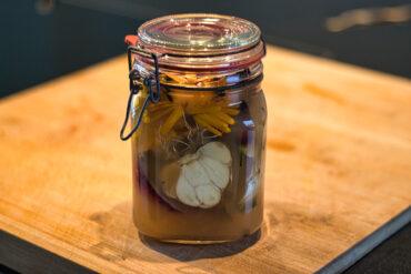 A glass jar with a metal clasp lid sits on a wooden surface, filled with pickling liquid, garlic, and herbs. Yellow flowers and a halved garlic bulb are visible inside the jar.