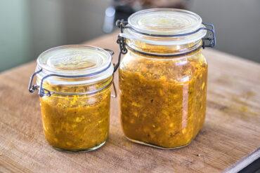 Two glass jars filled with a chunky yellow relish or chutney sit on a wooden surface. The jars have metal clasp lids and are of different sizes. The relish contains visible bits of vegetables and spices.